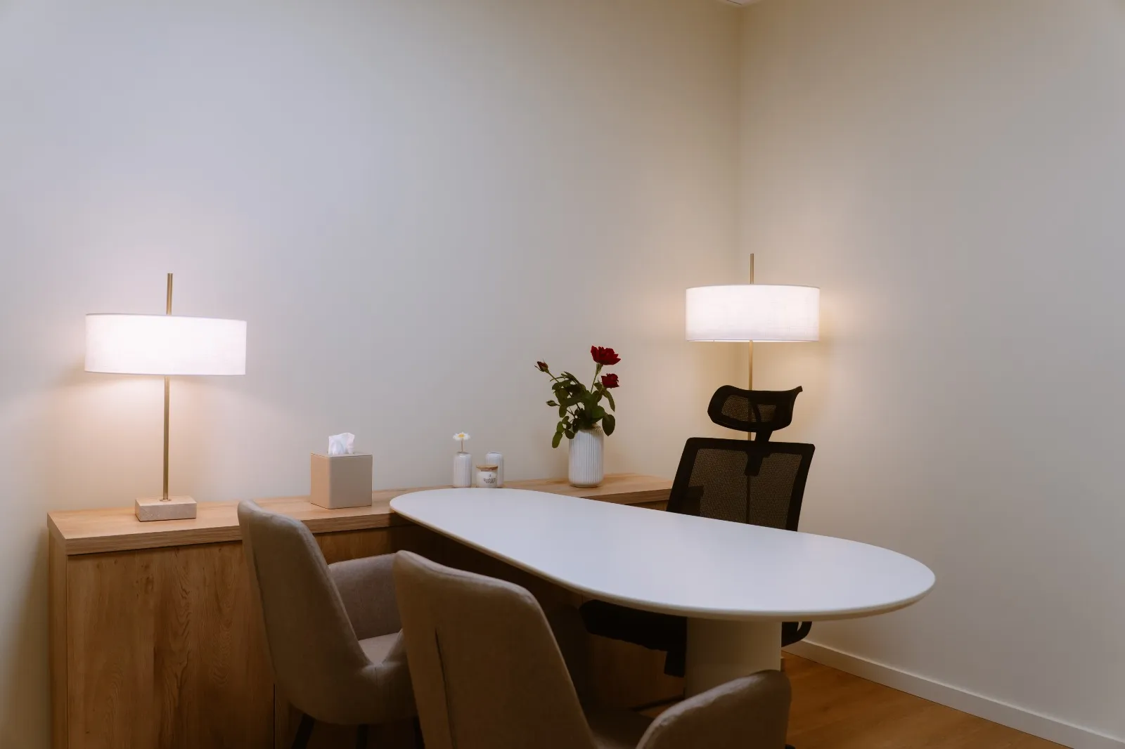 Sala de consulta com mesa branca, cadeiras estofadas, candeeiro e flores